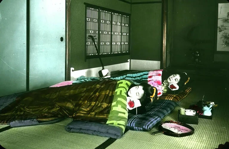 Interior shot of two geishas asleep in bedding on floor mats; musical instrument, fan and implements for tea ceremony nearby.