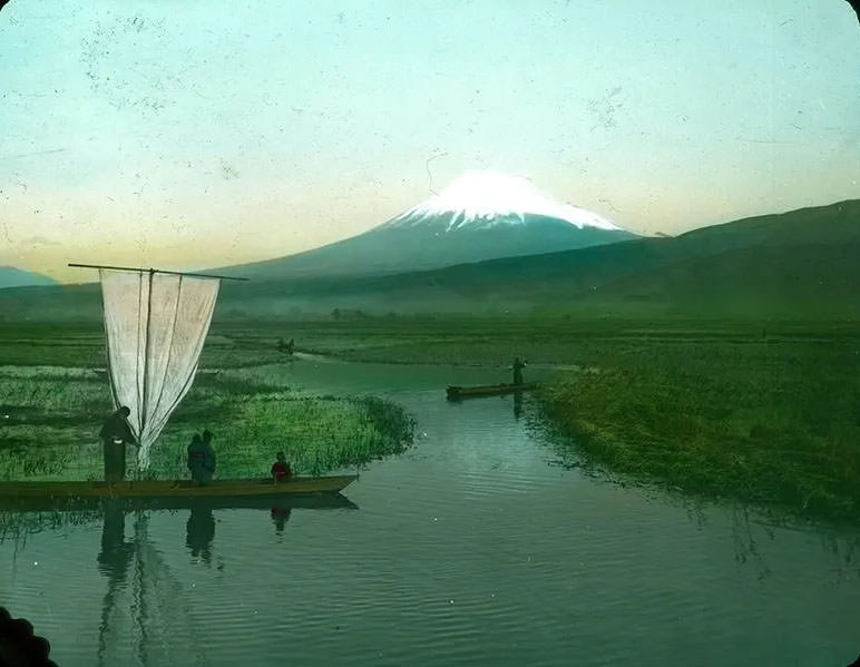 People in boats on waterways through rice fields; snow-covered mountain in background.
