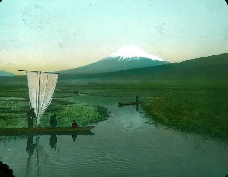 People in boats on waterways through rice fields; snow-covered mountain in background.