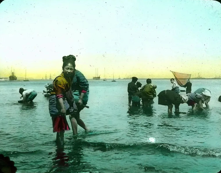 People wading in sea; fisher with hand net; boats in background.