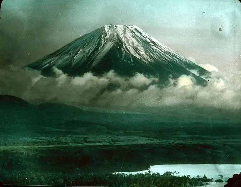 Snow-covered mountain (Mt. Fuji ) wreathed with clouds; low ground leading down to water in foreground.