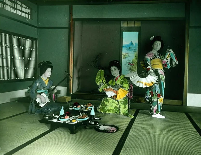 Three women with fans, musical instrument and a table set with food.