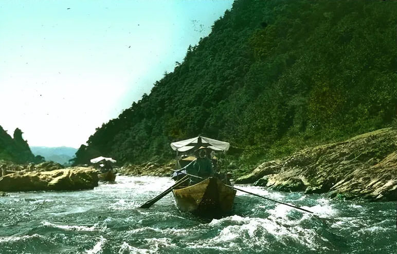 Two boats with cloth canopies being rowed along river; wooded mountain on one side; rushing water in foreground.