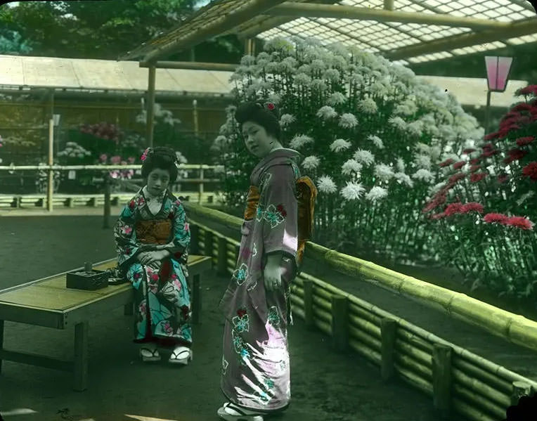 Two young women in Kimonos, one standing, one seated on wooden bench outside open greenhouses filled with blooming flowers.