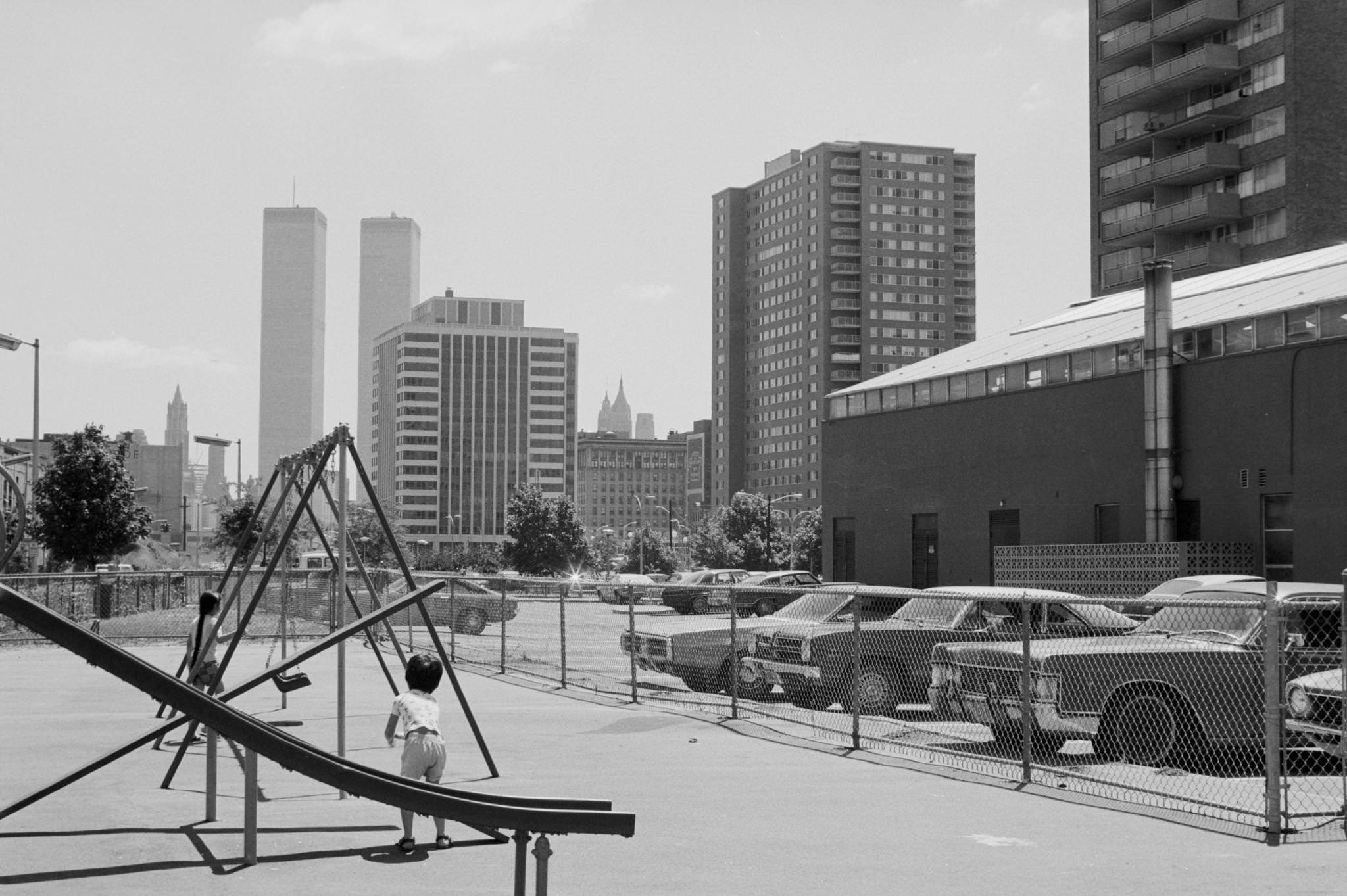 With the World Trade Center in the distance, view looking east at two children at play in a playground in a housing complex in downtown Jersey City, New Jersey, 1974.