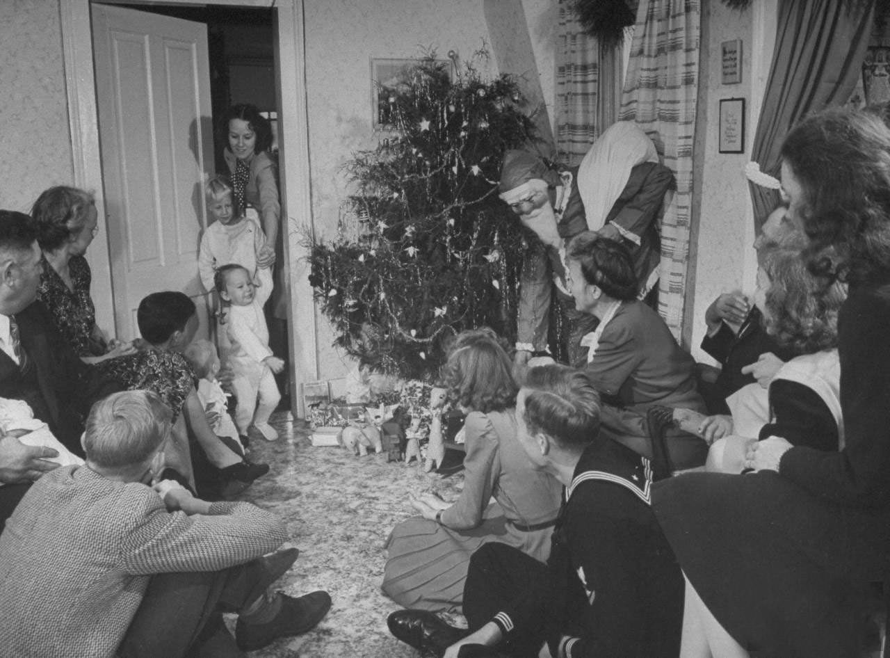 Adult members of farmer James Ferdinand Irwin's family gathered nr. tree watching his brother-in-law Fred Andrews (in Santa Claus costume) give presents to young family members at early Christmas family reunion marking safe return of sons from service in .WWII