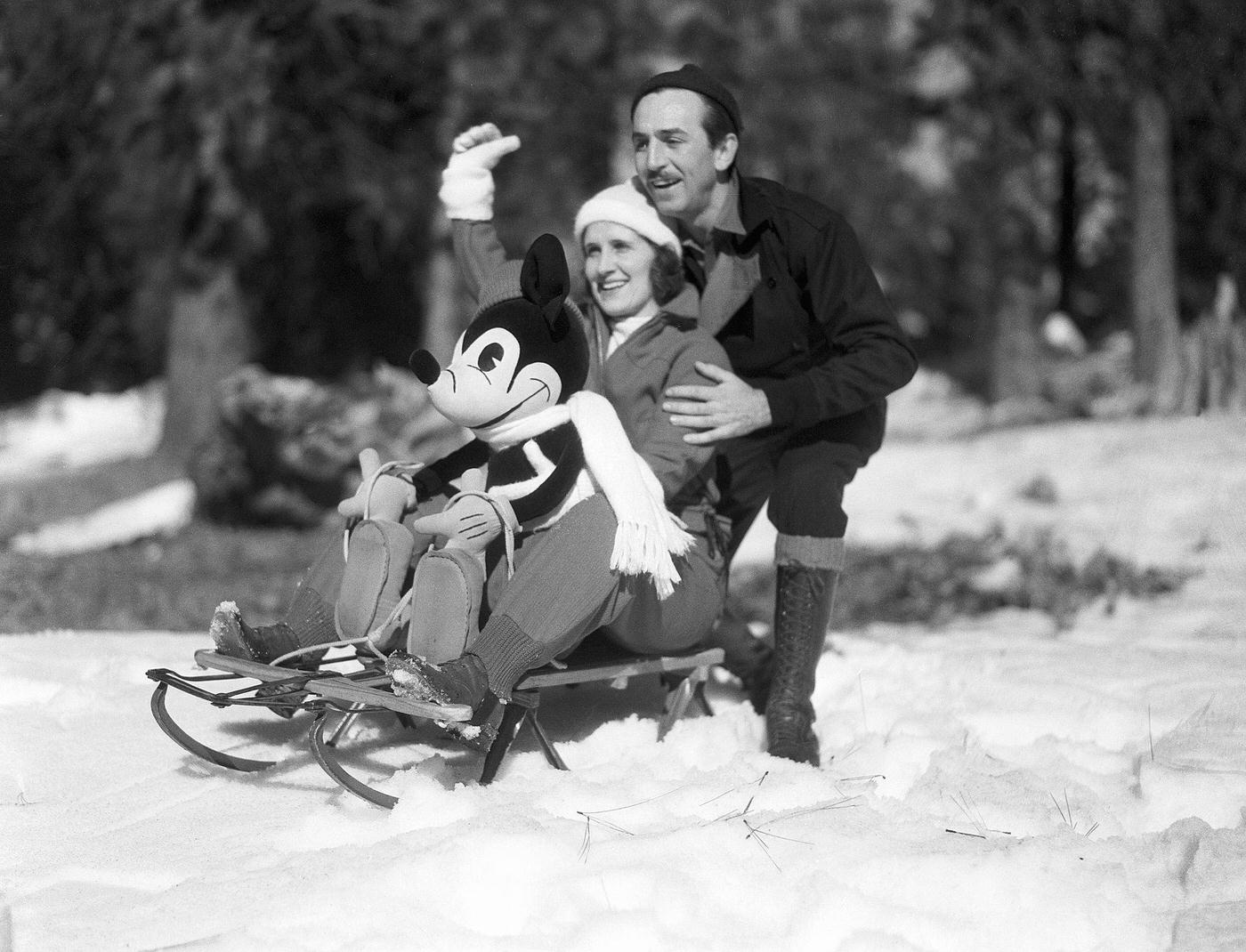 Mr. and Mrs. Walt Disney rest on a sled with a stuffed Mickey Mouse as they greet children to a snow party at the Lake Arrowhead resort area.