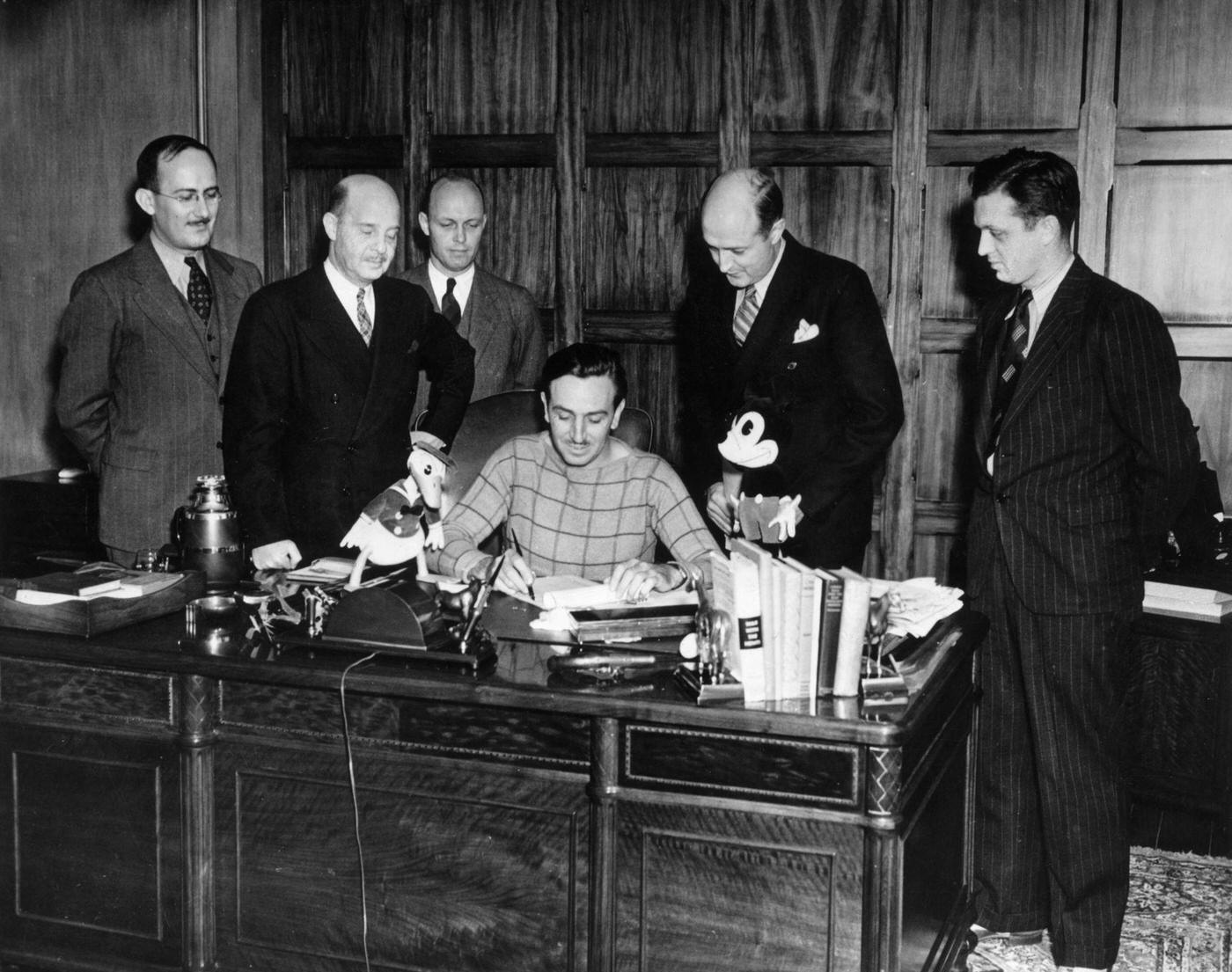 Walt Disney surrounded by a group of executives while seated at his desk in his office. Donald Duck and Mickey Mouse figures stand on the desk