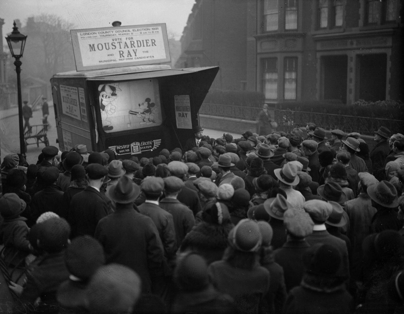 During an LCC (London County Council) election campaign run by Sir W Ray, Mickey Mouse is showing on a small screen in a city street, 1931