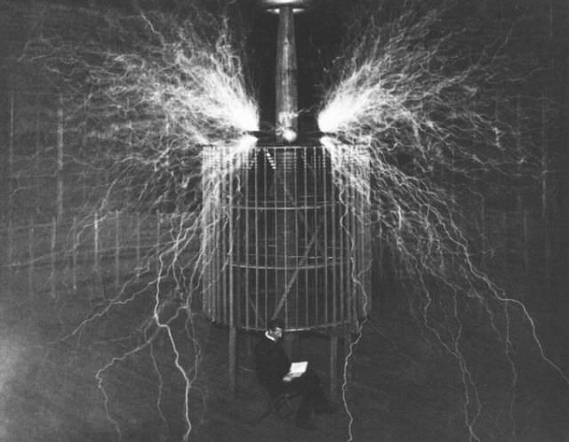 A glow of nitrogen fills the atmosphere. Tesla is photographed sitting in front of his generator. This photograph was taken in 1899.