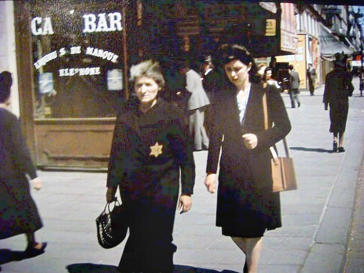 An elderly woman walks along the street wearing the yellow star that Nazis forced Jews to wear.