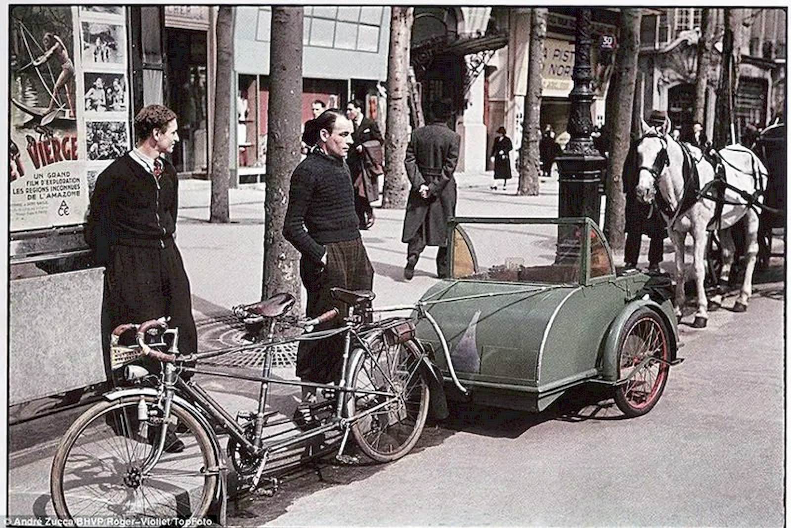 Two fashionably dressed young men stand by a tandem bicycle towing a carriage of sorts.