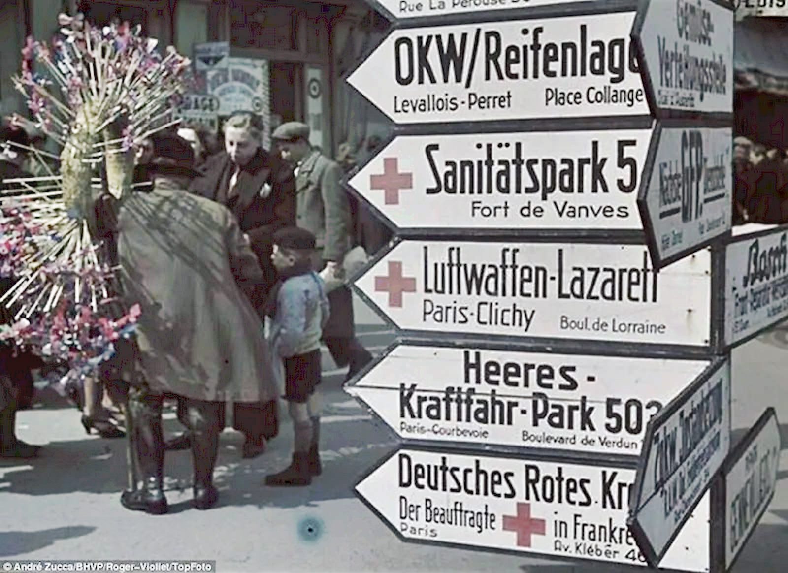 Street signs advertise locations of German facilities in Paris, with their French names written in smaller text beneath.