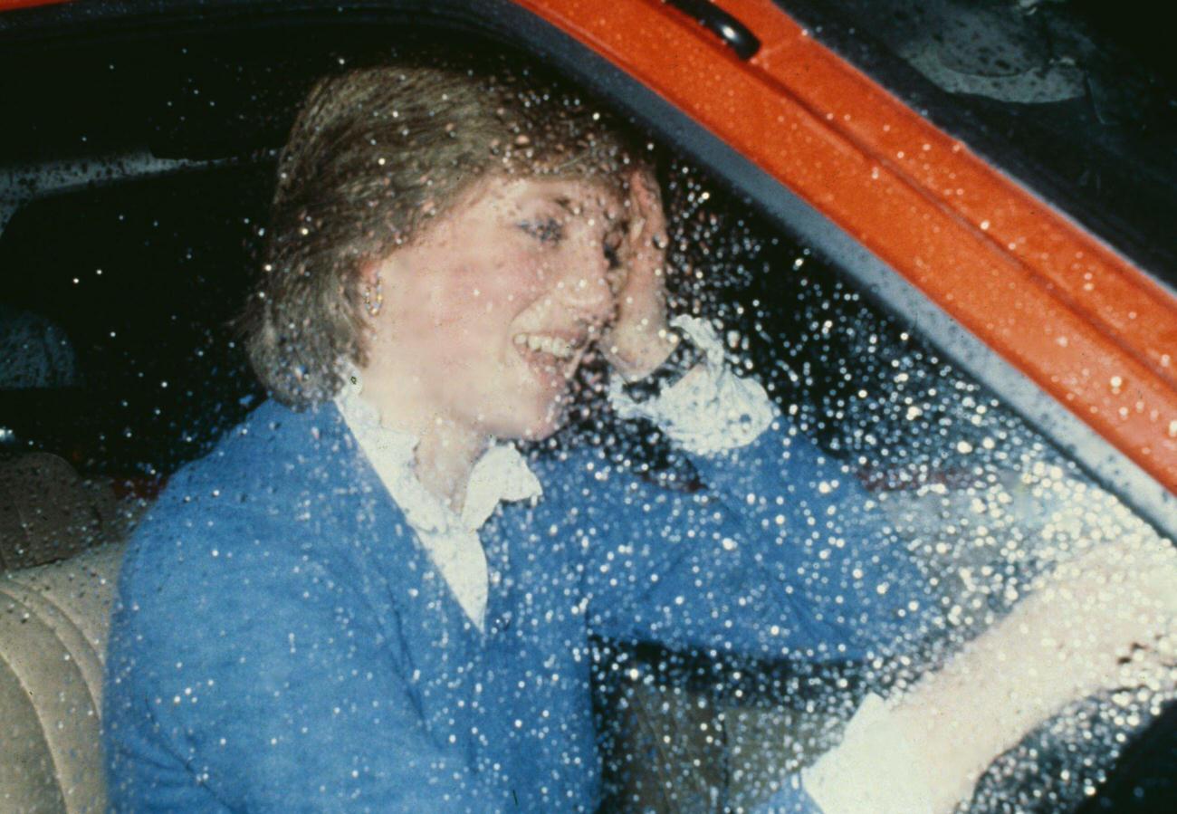 Lady Diana Spencer wearing a blue sweater over a white blouse, rain on the driver's seat window as she drives her Austin Metro car, 1980