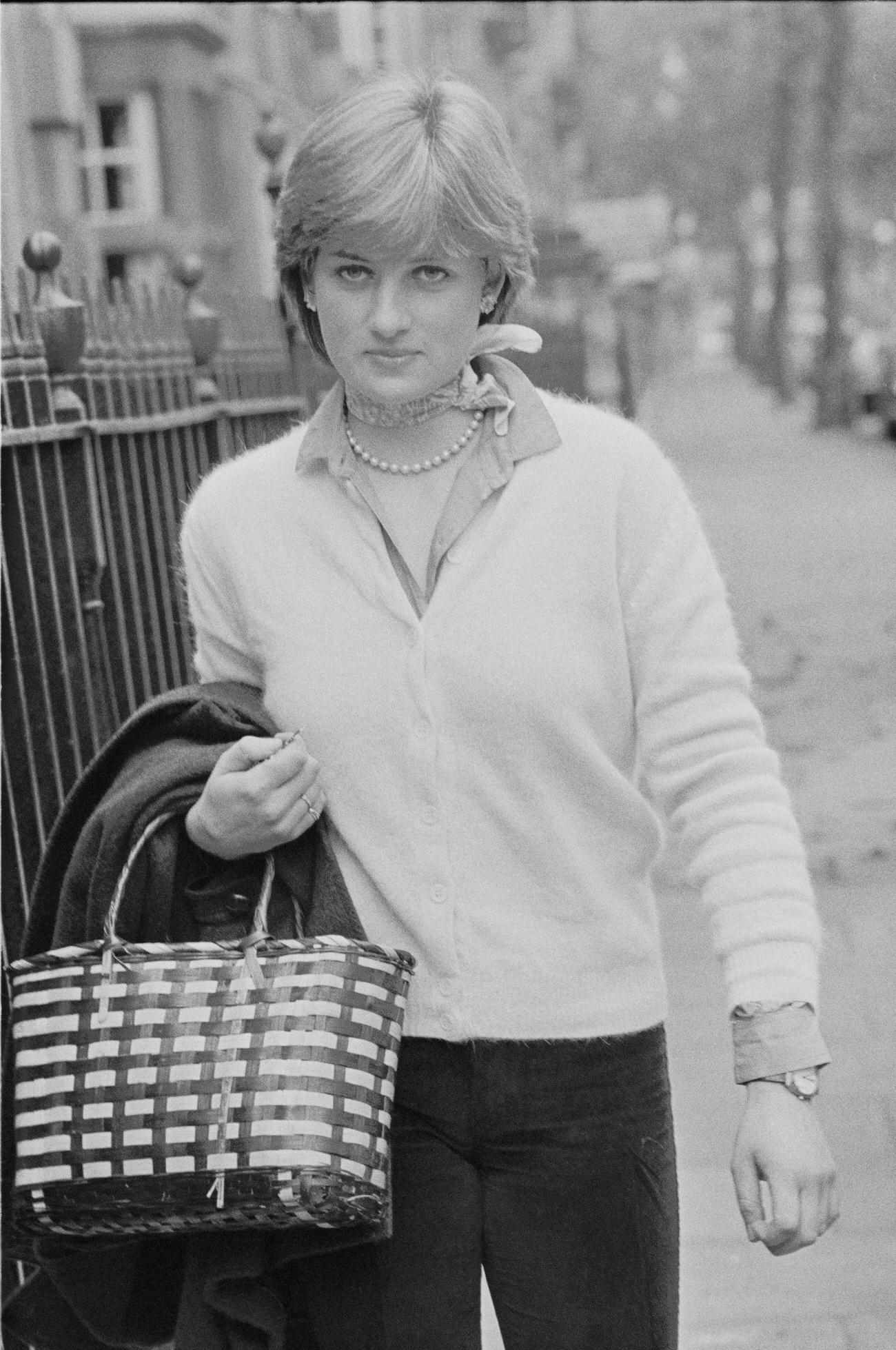 Lady Diana Spencer walking on a sidewalk holding her coat and handbag, she is wearing pale cardigan, shirt, neck-scarf and pearl necklace, London, 1980