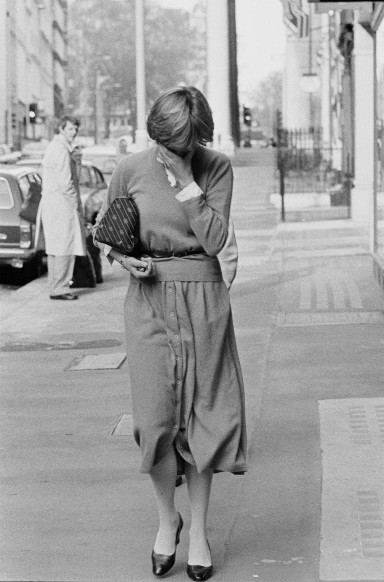 Lady Diana covers her face as she walk on a street in London, UK, 17th November 1980; she is wearing cardigan and midi skirts buttoned on front.
