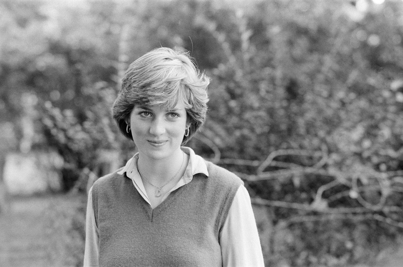 Lady Diana Spencer, wearing a floral pattern dress with a sweater draped over her shoulders, her head bowed with her left hand covering her face to avoid photographers, in St George's Square.