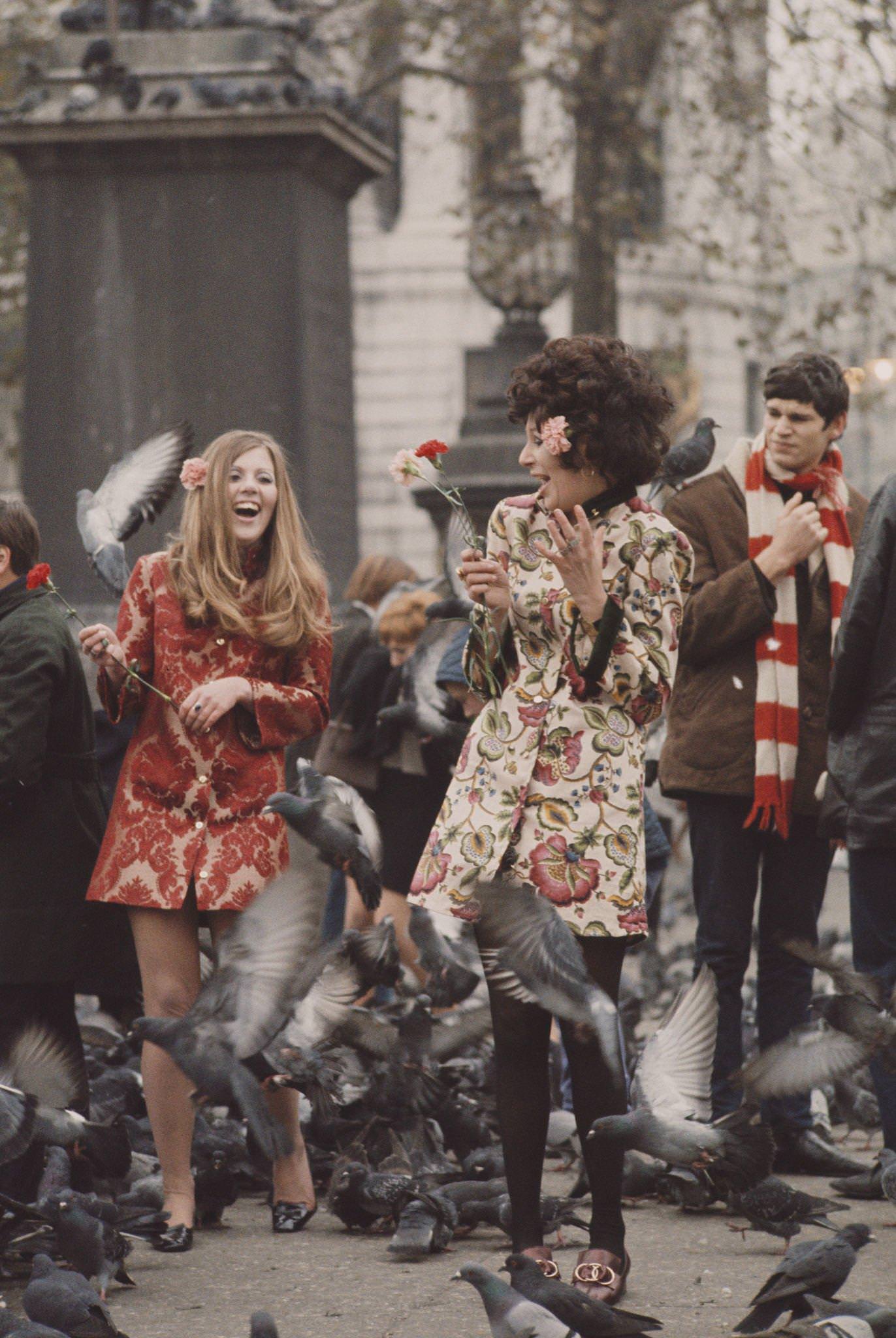 Two young girls dressed in short floral brocade-print coats stand holding flowers as pigeons fly around them in Trafalgar Square, London in November 1967.