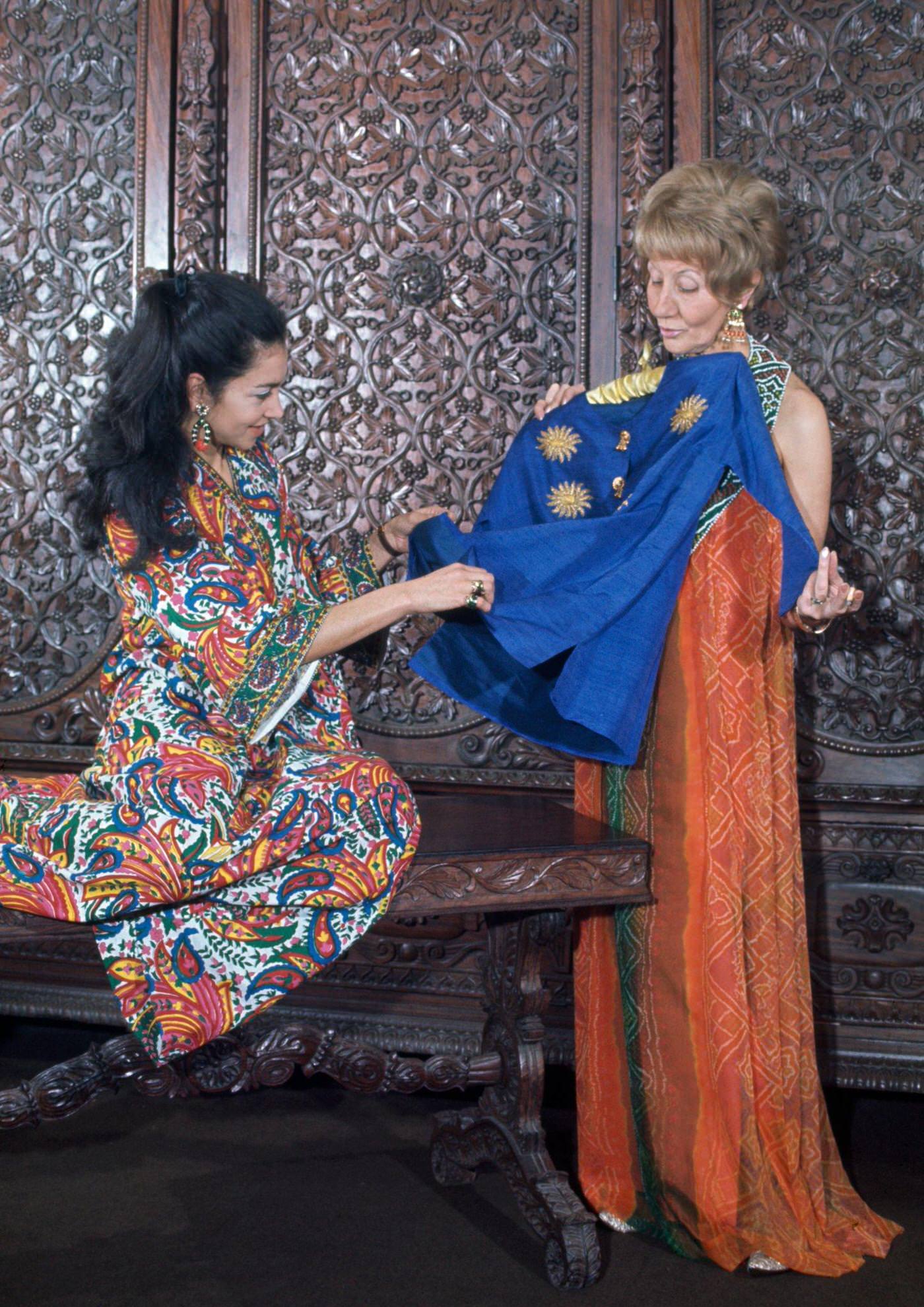 Two women in a hippie fashion boutique, one wears a long orange halter neck dress, the other wears a kaftan dress in a vibrant floral print, London, 1966.