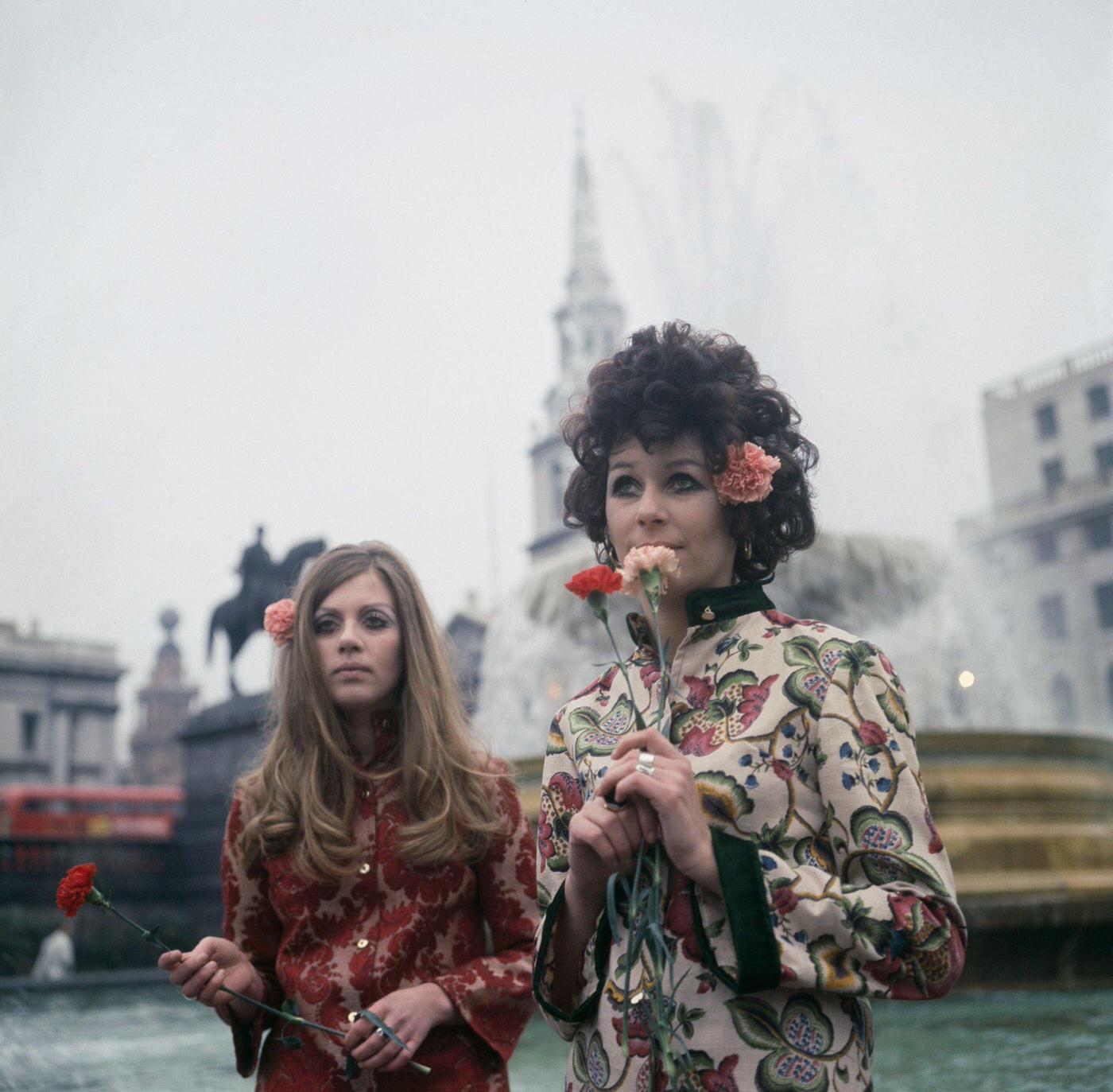 Two young girls dressed in floral print hippie style tunics stand holding flowers in Trafalgar Square, London in November 1967