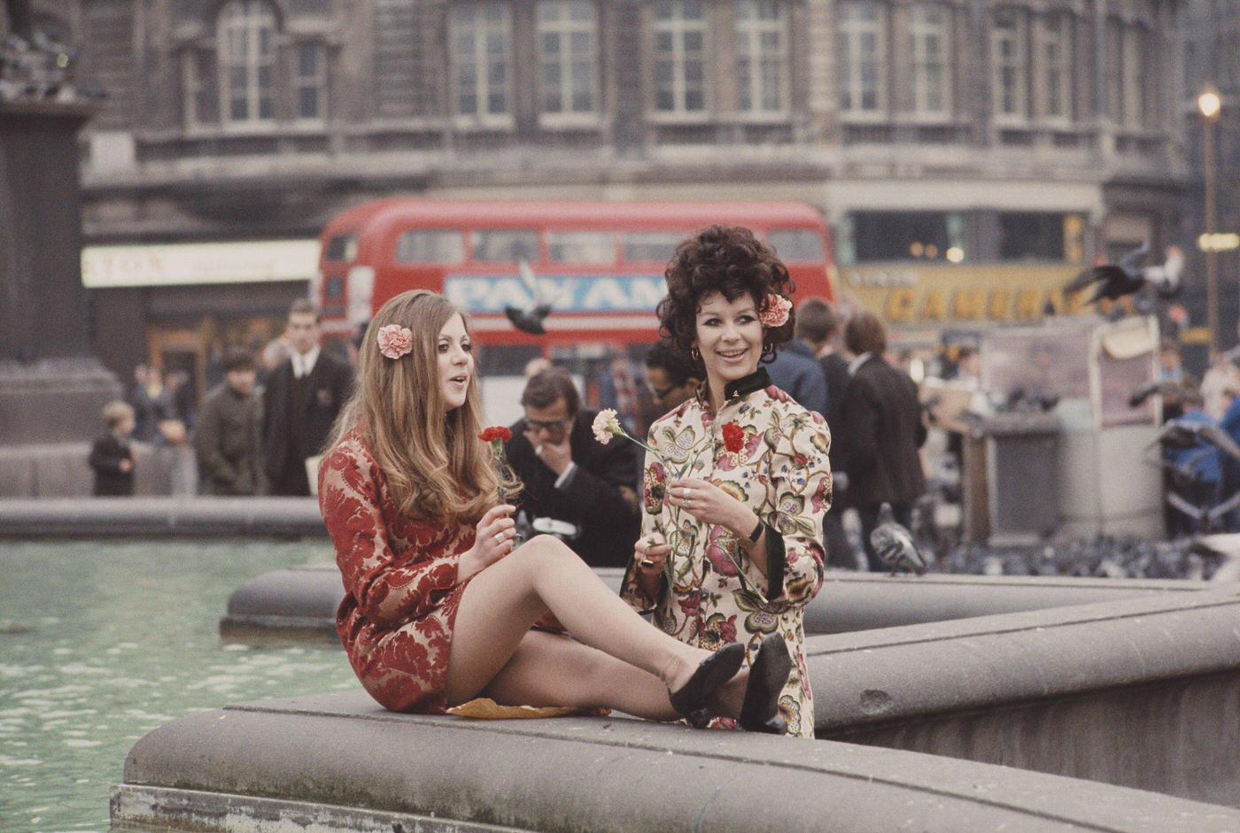 Two young girls dressed in floral print hippie style tunics stand holding flowers beside ornamental fountains in Trafalgar Square, London in November 1967
