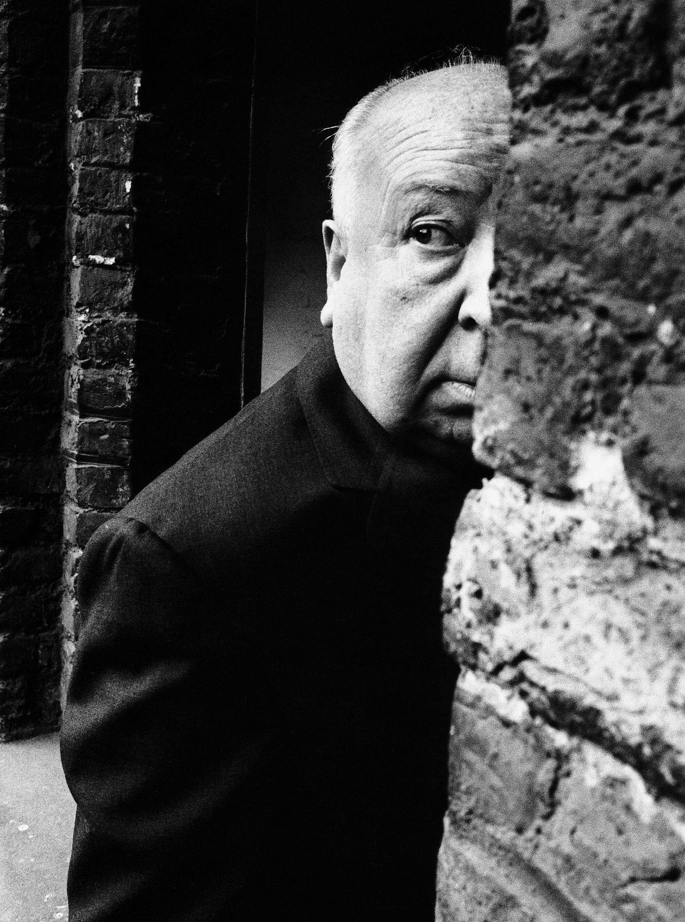 Alfred Hitchcock, looks as though someone might be following him as he disappears behind a wall.