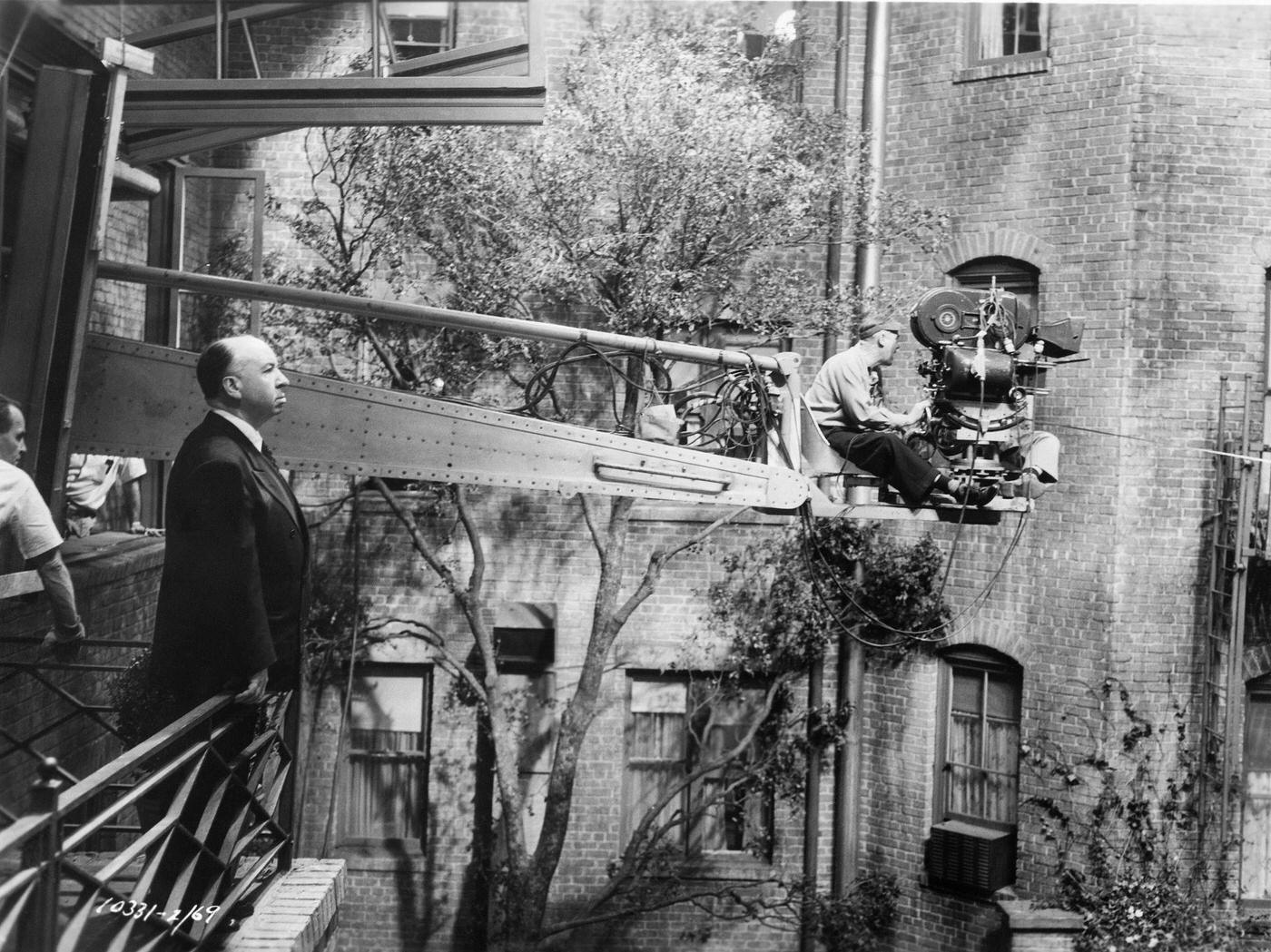 Alfred Hitchcock observes the action from a balcony as cameramen extended out on an "arm" film a scene on the set of "Rear Window" 1954.