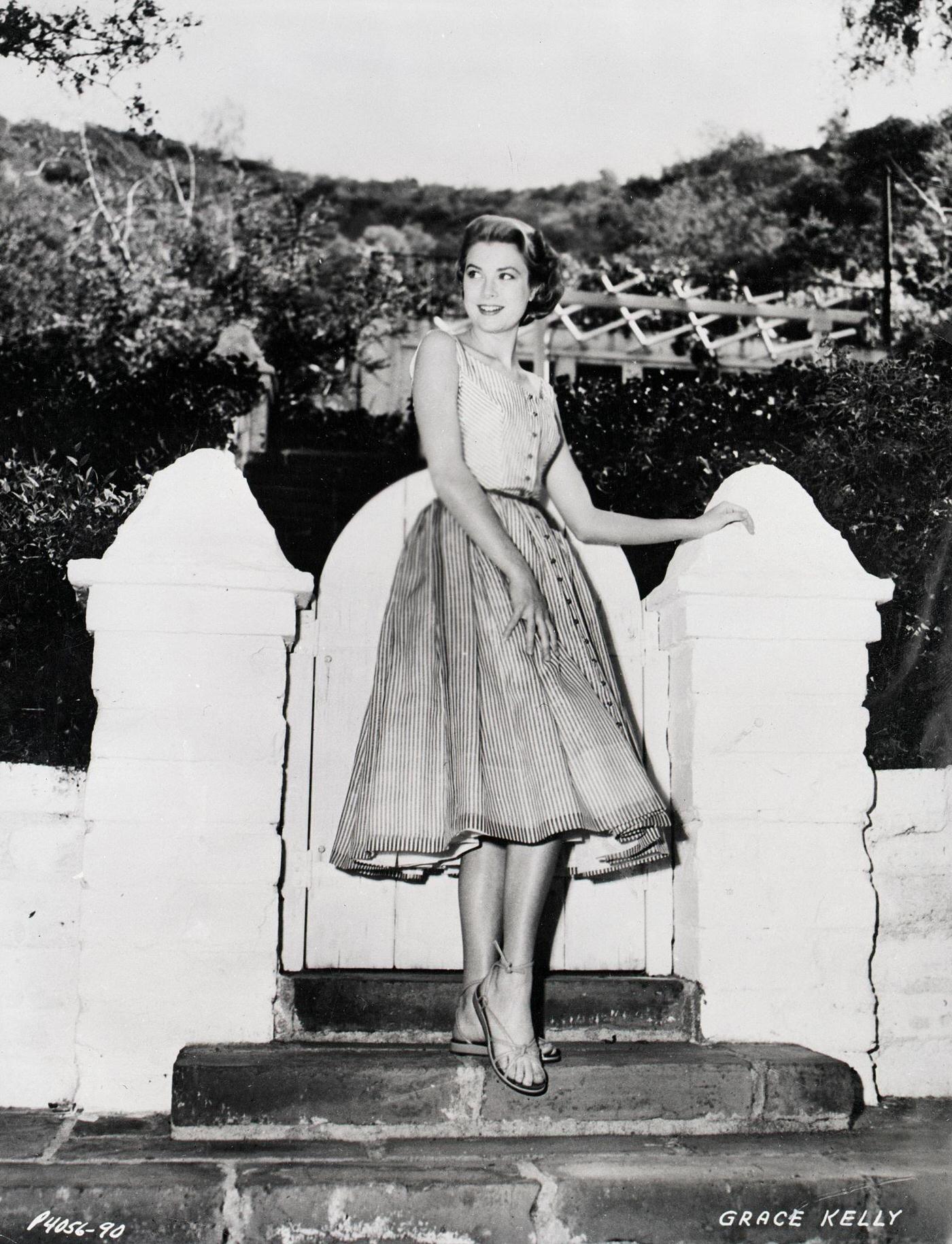 Grace Kelly models an Edith Head designer dress by a garden gate, 1954