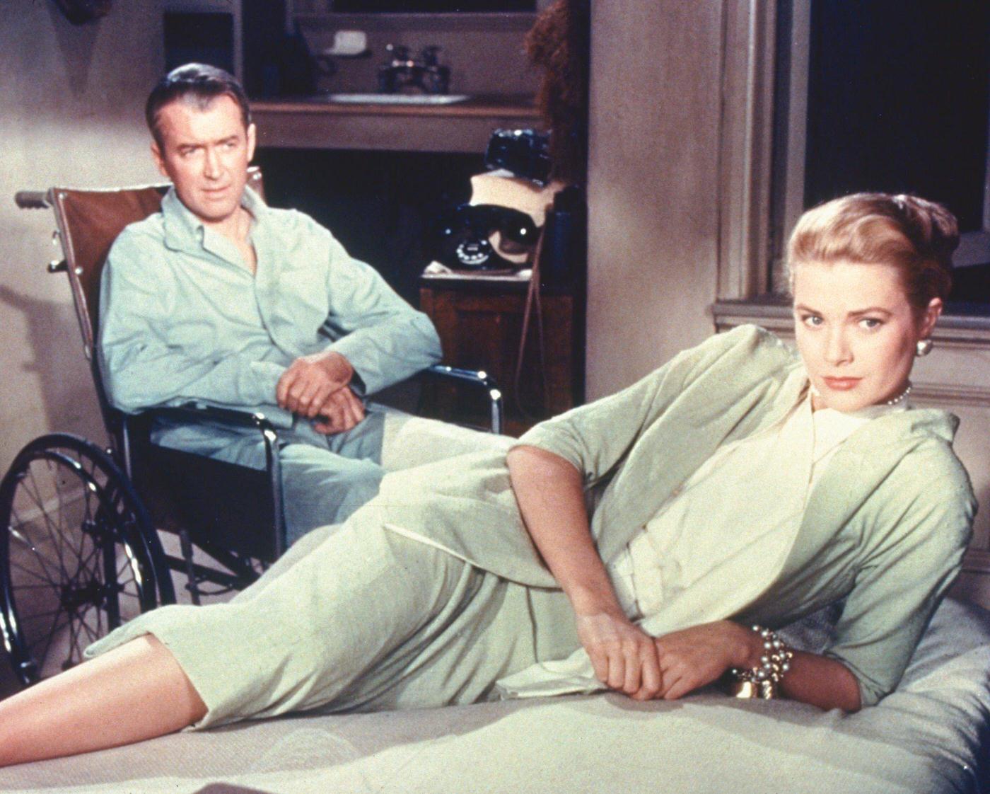 James Stewart wearing pyjamas as he sits in his wheelchair, while Grace Kelly lays on a bed in a publicity still issued for the film, 'Rear Window', 1954