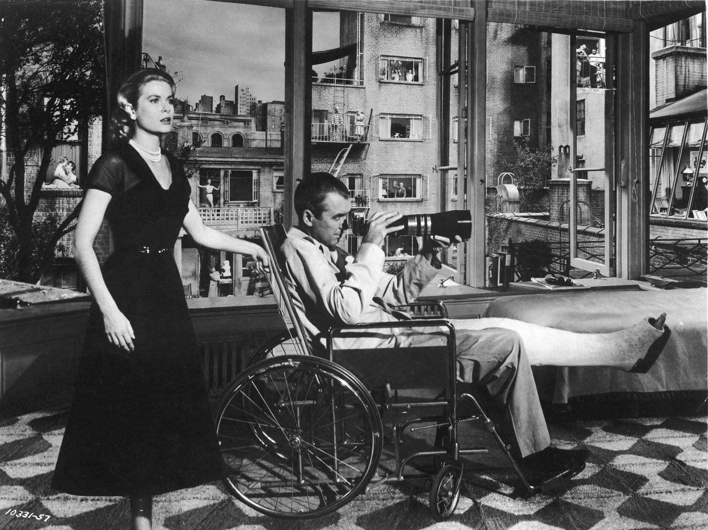 Grace Kelly stands behind actor James Stewart who sits in a wheelchair with his leg in cast,
