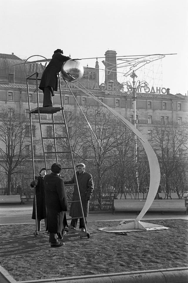 Four men installing a model of Sputnik 1 on an abstract sculpture in a public garden.