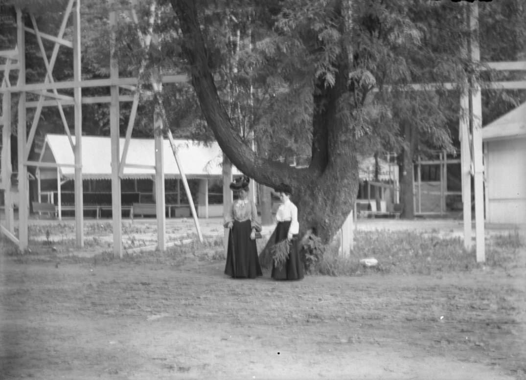Two women standing by a large, misshapen tree near a tall, wooden structure, 1900.