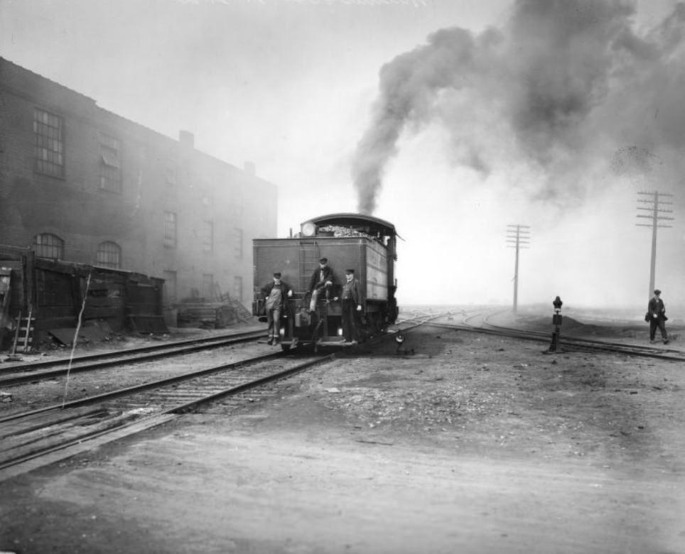 Rear of a railway locomotive travelling away from the camera. Trainmen and an engineer are on the back of the Tender.