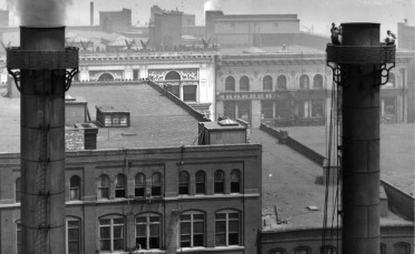 A pair of smokestacks, one of which is being worked on by a group of men near the top of the stack, 1900. The upper stories and roofs of 901 and 911 Washington Avenue are visible in the background.