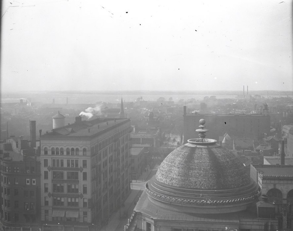 An aerial view of Saint Louis. The view spans across rooftops with smokestacks in the distance, and the river in the background, 1900