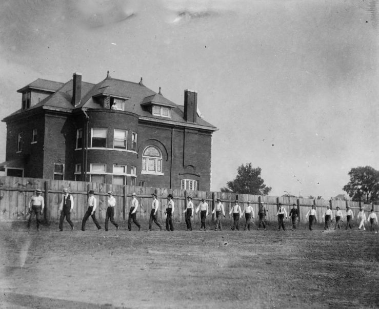 Several unidentified men in line along fence, 1900