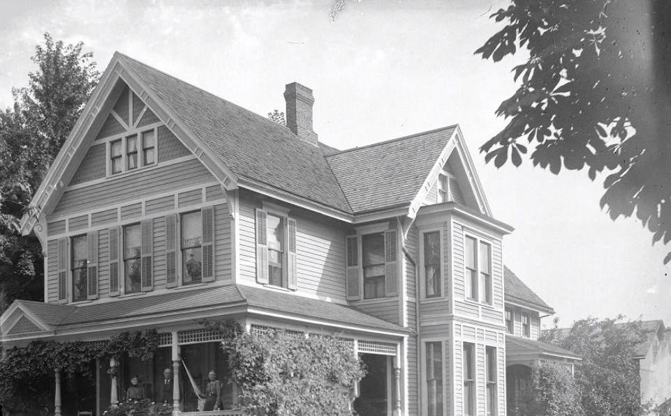A large house with four people on and around the porch facing the camera, 1900. Going towards the right of the photo, the neighborhood continues with vegetation grown houses.
