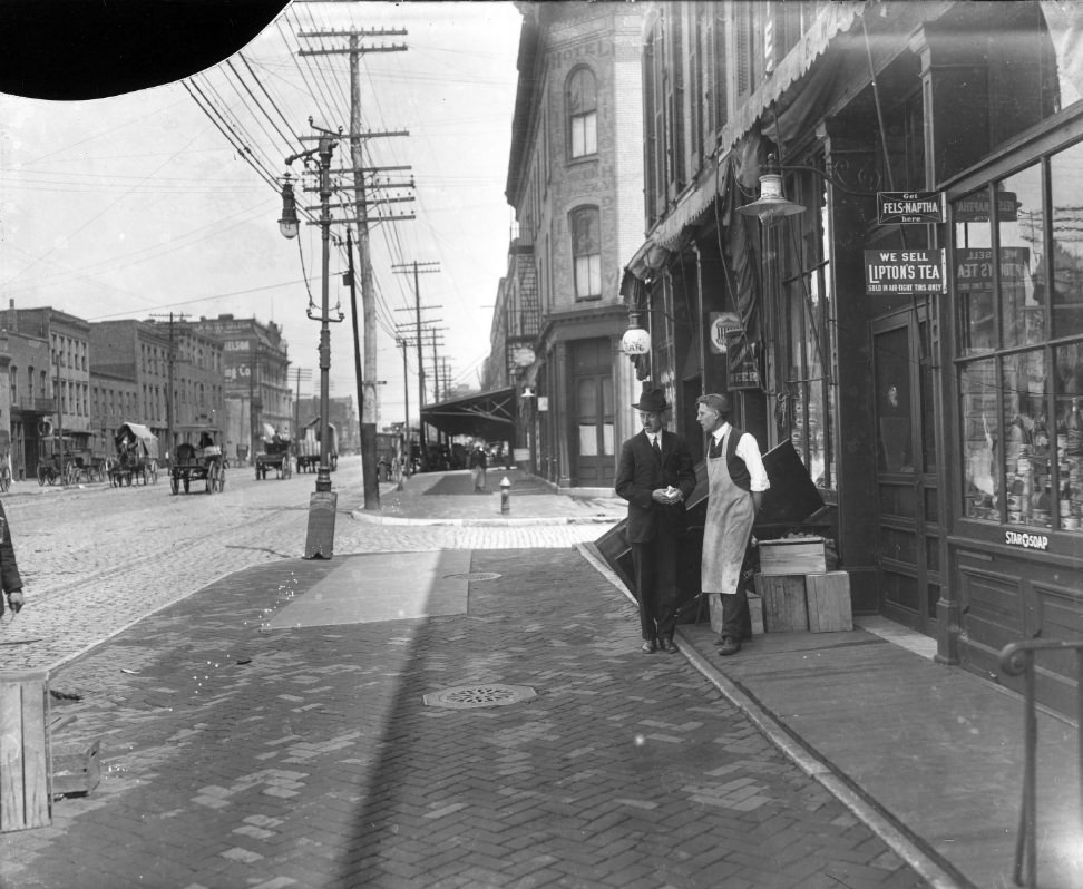 Two men standing on the sidewalk of a major street. One man appears to work at the grocery they are standing in front of, 1900