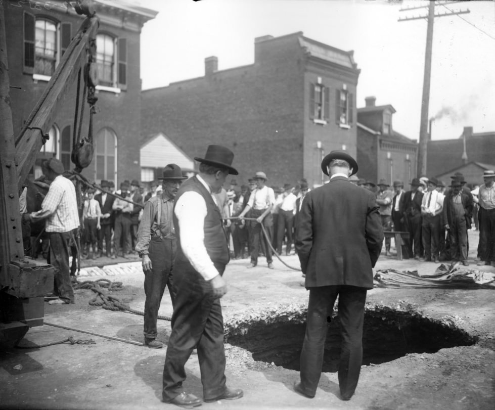 People standing around a large hole in a city street, 1900