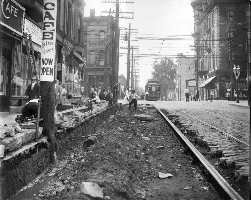 A section of cobblestones has been removed along a busy sidewalk, 1900