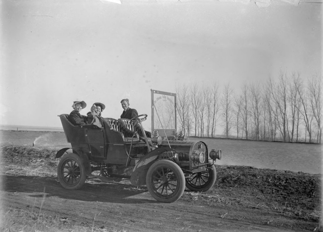 Three people in a company care parked by the river. Two women are sat in the back of the car, and a man is standing next to the car; all smiling.