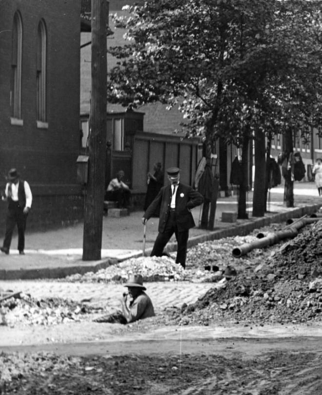 Men installing a pipe in a city street. One man is standing in a hole in the middle of the street and is smoking a pipe, 1900