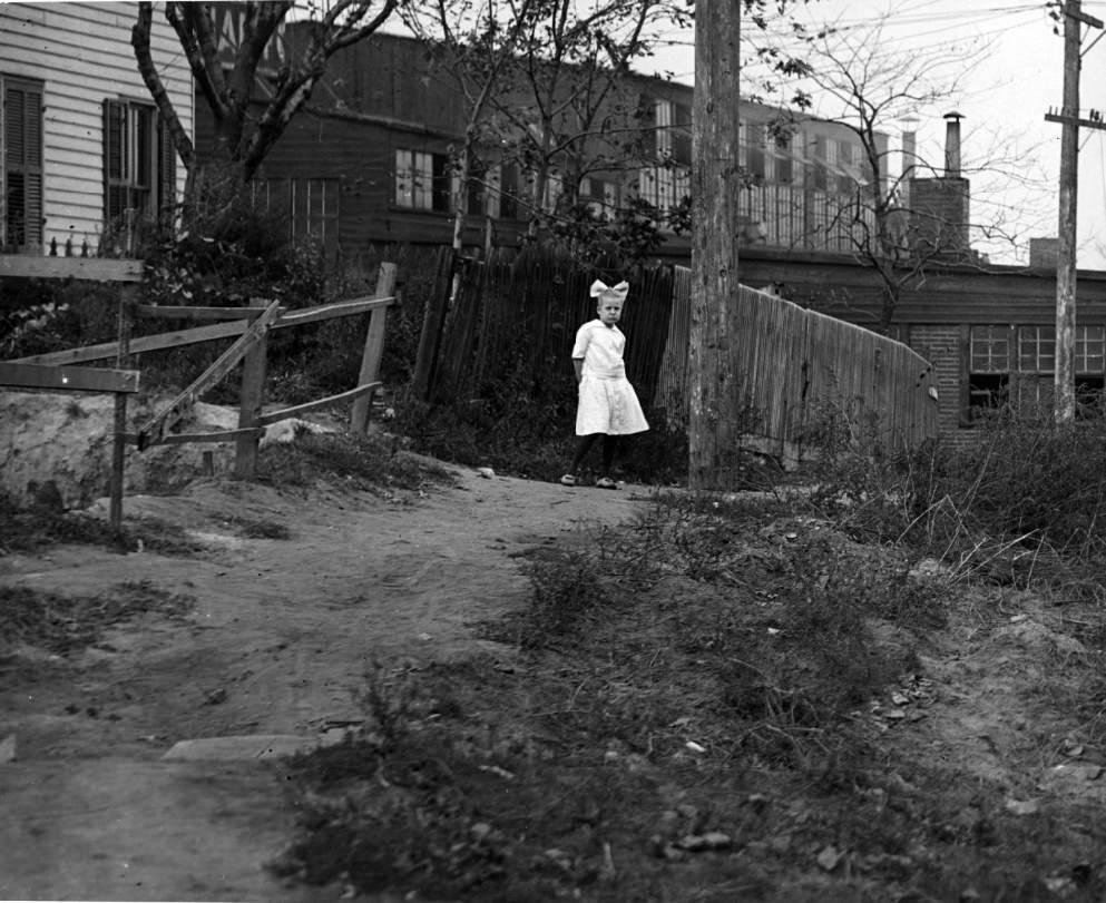 Girl on dirt path, 1900. An industrial warehouse is visible in the background.