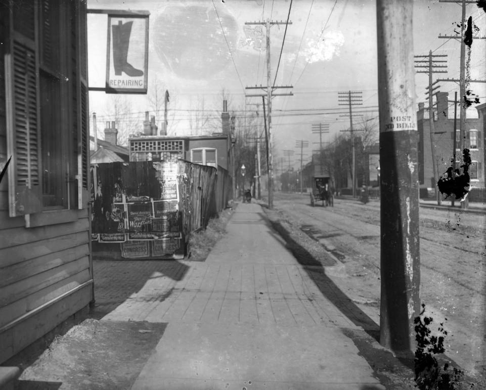 City sidewalk with wooden fence with posters plastered on it, 1900.