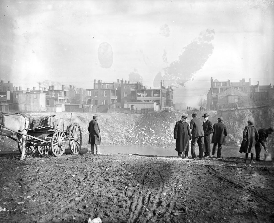 Large pit in the middle of an urban area that is filled with water, 1900.