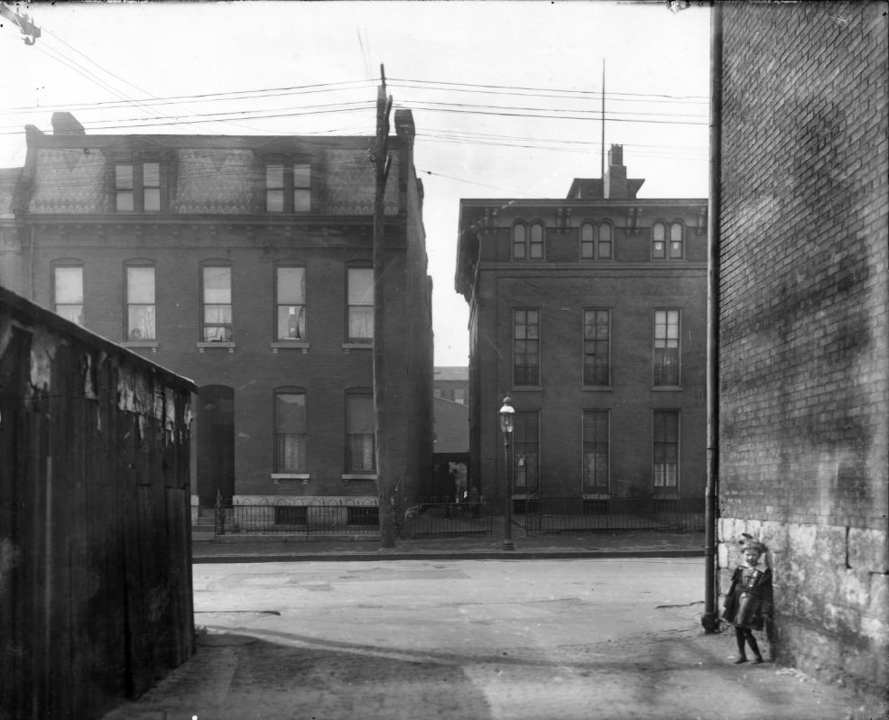 The front of two city houses side by side, 1900. Buildings in foreground partially obstructing the house fronts. Young child in alley in foreground.