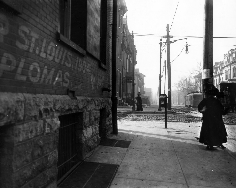 Street scene of St. Louis after during or after 1904. Two women on sidewalk. Trolley car on rails in the cobblestone street. Buildings with mansard roofs.