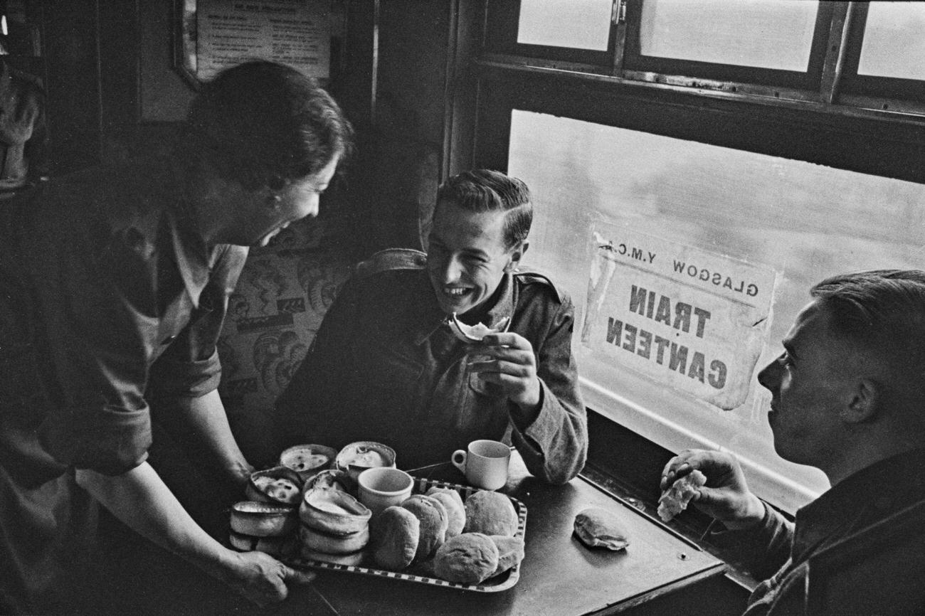 A female worker from the Young Men's Christian Association (YMCA) serves pies and rolls to British servicemen from their on-board canteen in a train carriage during a railway journey from Glasgow