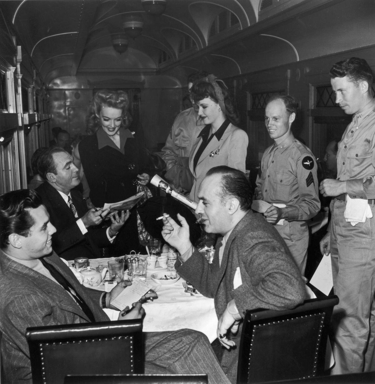 A group of actors chat around a table as three American servicemen look on, while traveling by train on the Hollywood Victory Caravan.