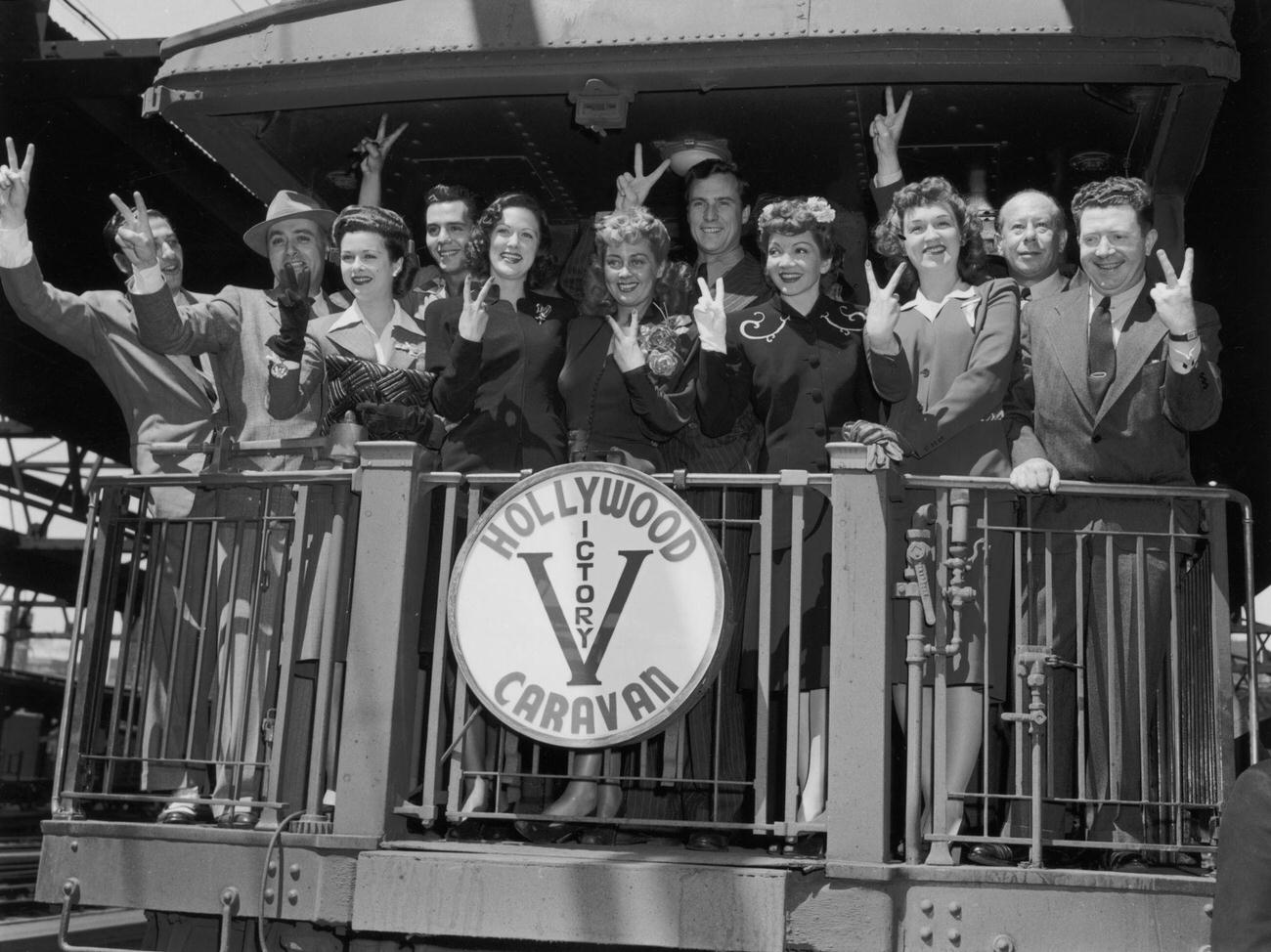 Members of the Hollywood Victory Caravan make a 'V' for 'Victory' sign while standing at the back of a railroad car.
